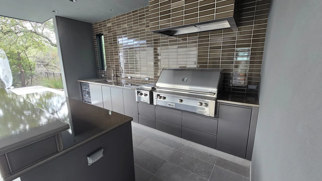 Outdoor kitchen with dark casework, a stainless steel grill and hood, a sink and a tiled backsplash.