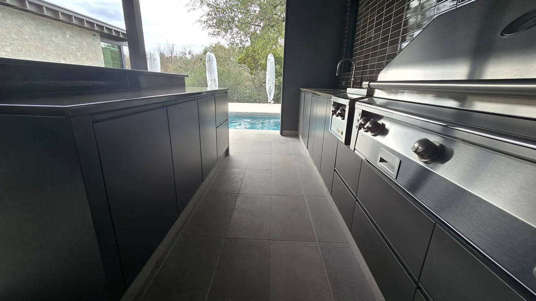 Dark outdoor kitchen cabinets and drawers alongside a stainless steel grill, with an island and pool beyond.